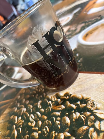 A close-up, angled shot shows a clear glass mug tilted to the left, filled with dark coffee. The mug has a decorative design that includes a heart shape and the words "Sweet Home" in gold script, along with feather-like patterns. The mug is positioned over a surface that appears to be a placemat or tablecloth printed with an image of roasted coffee beans. In the background, out of focus, are hints of other objects, possibly in a domestic setting. The overall scene is a still life focusing on a coffee mug and its thematic background. The location in the city of Ves', Russia, is not evident from the image.