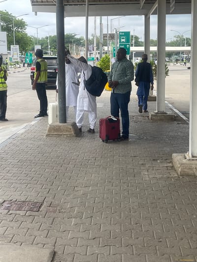 The image depicts an outdoor covered waiting area, likely at a transportation hub such as an airport or bus terminal in Gwaska, Nigeria, during daylight hours. The sky appears bright and overcast.
In the foreground, the ground is paved with grey interlocking concrete blocks. Several individuals are standing or waiting. Towards the center-left, a man wearing a green and white plaid shirt and dark blue jeans stands beside a red rolling suitcase, holding a mobile phone. Behind him, another man in a blue tunic and cap is visible. Further to the left, a man dressed in white traditional attire (possibly a thobe or kaftan) with a black backpack stands near a white rolling suitcase. He appears to be looking down or speaking to someone. Two individuals wearing yellow high-visibility vests and dark trousers, possibly security or staff, are positioned on the far left and right side of the foreground and midground.
The area is covered by an overhead structure composed of concrete and metal beams, providing shade. In the background, a roadway is visible with several parked and moving vehicles, including a white van. Greenery, consisting of trees and shrubs, dots the landscape beyond the road.
Notable text includes a green road sign displaying a bus icon next to "Bus" with a left-pointing arrow, and "Hall" with an upward-pointing arrow. Another yellow sign in the distance is partially visible, showing "Your goods. Your smile." and "Tatum Bank". A manhole cover embedded in the pavement in the midground has the text "FEDERAL ROADS" partially visible. The overall scene suggests a routine day at a busy public transport facility.