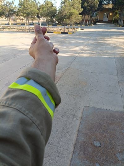 A close-up, first-person perspective shot shows a person's hand making a heart gesture, fingers forming a circle, in front of a paved area. The person is wearing a tan jacket with reflective yellow and grey stripes around the cuff. The background features a row of trees and a building. The paved ground is cracked and shows visible expansion joints. The scene appears to be an outdoor area, possibly a courtyard or walkway. Based on the context provided, the location is the city of Balkanabat City, Turkmenistan.