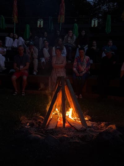 An outdoor evening scene shows a group of people sitting on tiered wooden benches, facing a campfire. The setting appears to be a campground or outdoor gathering area in Ratnieki, Latvia, as indicated by the distinctive lanterns hanging above the seating area. The campfire, in the foreground, is actively burning, casting a warm glow on the immediate surroundings. The people are casually dressed, some appearing to be young adults and others older. They are seated in various postures, some looking towards the fire, others conversing or resting. The background is dark, with trees visible against the twilight sky. Decorative elements, possibly flags or streamers, are also visible hanging from poles above the benches. The overall atmosphere suggests a relaxed communal gathering around a campfire.