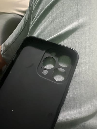 The image depicts a black smartphone case resting on a person's leg in Karnalovo, Bulgaria. The case, designed for a multi-lens camera phone, features four large circular cutouts and one smaller circular cutout on its rear surface. Minor scuffs are visible on the matte black finish of the case. The leg is clothed in a light green or teal-colored fabric, likely trousers, exhibiting folds and a textured surface. In the upper-left background, a portion of a lighter-colored, possibly off-white, upholstered surface is visible. No other individuals or specific environmental details beyond the immediate area are discernible.
