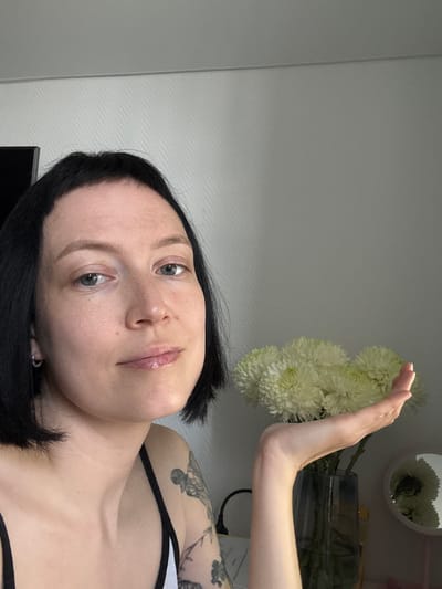 A close-up, indoor, eye-level shot shows a person with short black hair and light-colored eyes. They have a subtle smile and are looking towards the camera. Their left arm is raised, palm open, towards a bouquet of white chrysanthemums in a dark blue vase. Tattoos are visible on their upper left arm and shoulder. A circular mirror with a reflection of flowers is positioned to the right of the vase. The background consists of a textured, light-colored wall and a hint of a television screen. The overall lighting is soft and natural. The image does not provide any information about the specific location in the city of Боголюбово, Russia.
