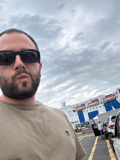 A medium-range selfie shot shows a man with a beard and sunglasses, wearing a beige shirt. He is positioned on a surface marked with yellow lines, next to several cars and trucks in a row. Behind the vehicles is a large ferry with a blue and white exterior. The sky is overcast with cloud cover. The photo was taken in Civitavecchia, Italy.