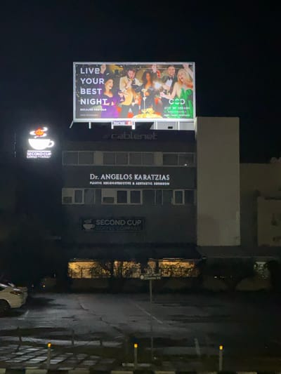 The image depicts a nighttime street scene in Engomi̇ Lefkoşa, Cyprus, featuring commercial establishments illuminated against a dark sky. The ground, likely a parking area, appears wet, suggesting recent rain.

A prominent dark grey building stands centrally, displaying two main signs. One reads "Dr. ANGELOS KARATZIAS PLASTIC RECONSTRUCTIVE & AESTHETIC SURGEON," and below it, another sign identifies "SECOND CUP COFFEE COMPANY." The building has multiple rectangular windows.

To the left of the building, a large, brightly lit billboard showcases a festive scene with a group of people celebrating, appearing joyful amidst confetti or streamers. The billboard features the prominent text "LIVE YOUR BEST NIGHT BECAUSE YOU CAN," along with "NICHE," "cablenet," and "CCD CITY OF DREAMS."

A smaller, ground-level illuminated sign further to the left also displays the "SECOND CUP COFFEE COMPANY" logo and name. A sliver of a vehicle is visible at the bottom, and striped bollards or barriers line the right side of the frame.