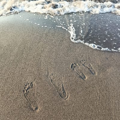 The image shows a sandy beach with three sets of bare footprints in the sand. The footprints are all facing the same direction. The image is taken from a high angle, looking down at the beach. The beach is located at the coordinates -7.71 latitude and 114.118 longitude, which is in Arjasa, Indonesia. The waterline of the ocean is visible at the top of the image.  The water has receded leaving behind a line of white foam.  The footprints are likely made by people who were walking along the beach.