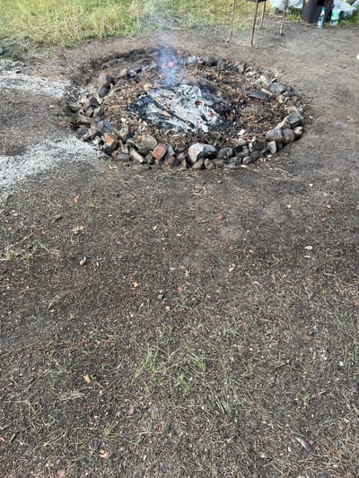 An outdoor, top-down view captures a fire pit constructed from stones and bricks in a campsite. The pit contains smoldering embers and ash, with wisps of smoke rising. Surrounding the fire pit is a mixture of dirt, sparse grass, pine needles, and small pebbles. In the upper right background, a metal grill, some plastic bags, and water bottles are visible. The ground is uneven and appears to be a natural, undeveloped area. The location is identified as Дражнаўскі сельскі Савет, Belarus.