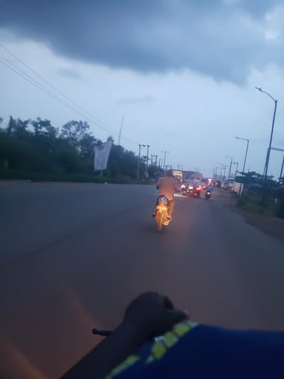 The image depicts a nighttime street scene, likely taken from a moving vehicle in Ida, Nigeria. A motorcyclist is visible ahead, followed by other vehicles with headlights illuminated. Streetlights line the road, and there are trees along the roadside, with a white flag visible on the left. The sky is overcast. In the foreground, part of the handlebar and sleeve of the photographer is visible.