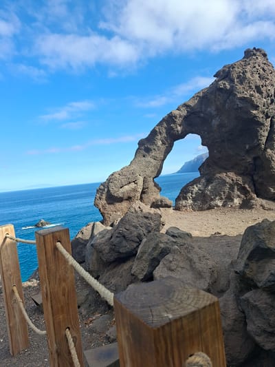 This is an eye-level, medium shot of the natural rock archway known as "El Dedo de Dios" (The Finger of God) on the coast of Santiago del Teide, Spain. The archway, a prominent geological formation, is composed of dark, rugged volcanic rock. It frames a view of the vast blue Atlantic Ocean, with a distant coastline visible on the horizon. The sky above is a clear, bright blue, dotted with wispy white clouds. In the foreground, a wooden railing with thick rope serves as a boundary, guiding visitors along the cliff edge. The ground is covered in dark volcanic gravel and rocks. The scene captures the raw beauty of the natural landscape, with the archway standing as a testament to the power of erosion.