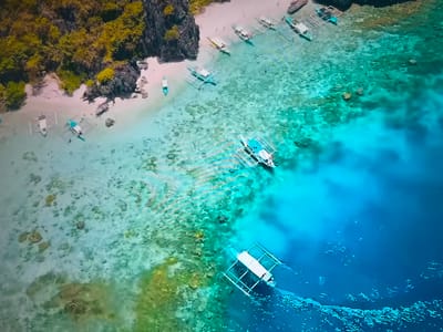 The image is an aerial shot of a tropical beach. A small island covered in vegetation is on the left side of the image. There is a sandy beach with several boats docked along the shoreline. The water transitions from shallow, clear turquoise near the beach to a deeper blue further out. Multiple boats with outriggers are visible in the water. The seabed is visible in the shallow areas, showing light-colored sand and rocks. The location is not in Basingstoke and Deane, United Kingdom; it is a tropical coastal environment.