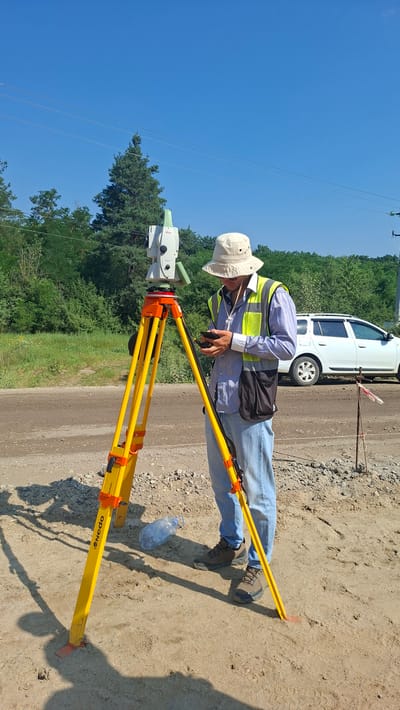 A person is standing in a sandy, outdoor area, operating surveying equipment. The person is wearing a light-colored bucket hat, sunglasses, a blue and white striped shirt, a yellow and grey high-visibility vest, and light blue jeans. They are holding a dark-colored mobile device. In front of the person is a tripod with a surveying instrument, a total station, mounted on top. The tripod is yellow with orange connectors and has a brand name "Nedo" visible on one of its legs. Behind the person, a white SUV is parked near a dirt road. The background consists of a line of trees and a clear blue sky. The environment appears to be a construction or surveying site. This scene is located in Hînceşti, Moldova.