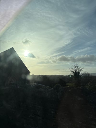 The image portrays a rural landscape in Snailbeach, United Kingdom, under a partly cloudy sky, indicative of either morning or late afternoon lighting conditions. A dark, stone-built structure with a prominent gabled roof and an arched entrance or window is heavily silhouetted in the left mid-ground, with a single-story extension, featuring a lighter roof, visible to its right. A large, bare-branched deciduous tree, also silhouetted against the bright horizon, occupies the right mid-ground. The background consists of undulating hills covered by sparse vegetation and distant treelines, with glimpses of green fields. The sun, partially obscured by clouds, is positioned in the upper left quadrant, producing significant backlighting, lens flare, and a prominent rainbow-like arc artifact in the upper left corner of the frame. The immediate foreground is rendered in deep shadow, appearing as an indistinct, dark mass of terrain. No individuals, explicit actions, or interactions are visible within the scene.