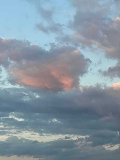 The media file displays a sky filled with a diverse array of clouds, characteristic of either a sunrise or sunset. The clouds exhibit a varied palette, predominantly featuring shades of grey and muted blue, with striking patches of soft pink and warm orange, particularly noticeable in the center and upper-left of the original image. These warmer tones suggest direct or indirect illumination from a low sun angle.
Interspersed among the denser cloud formations are areas of lighter, clear blue sky, indicating breaks in the cloud cover. Some clouds appear wispy and ethereal, while others are more voluminous and opaque, creating depth and texture in the scene. The lighting is soft and diffused, subtly highlighting the edges and contours of some clouds with pale, sometimes white, accents.
No people, objects, or specific activities are visible within the frame, which solely captures the atmospheric conditions. There is no visible text. The scene conveys a sense of natural beauty and tranquility. Based on the provided context, the image was taken in Zaķumuiža, Latvia.