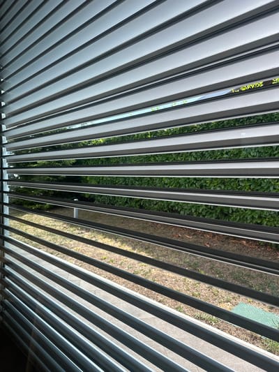 The image shows a view through horizontal blinds, likely in a residential setting. The blinds are light grey with darker grey trim. Through the slats, a green hedge or privacy fence is visible, beyond which is a patch of dry grass and some paving stones. The sky is visible in some gaps between the slats, indicating it is daytime. There are no people or animals visible in the image. The scene is devoid of specific identifying features that would place it in Trieste, Italy.