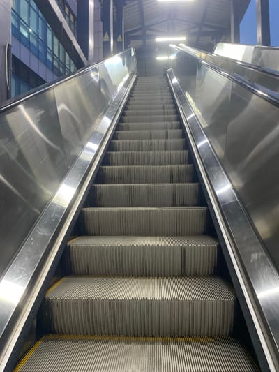 The image shows an upward-facing perspective of a stainless steel escalator at the St. Thomas Mount Cantonment railway station in Chennai, India. The escalator is clean and reflects the bright overhead lighting. The surrounding environment includes the metallic structure of the escalator, with its ribbed steps and brushed metal sides. In the background, a modern building with blue-tinted glass windows is visible, suggesting the station is integrated with urban infrastructure. The scene is devoid of people, presenting a quiet and empty state of the transit area. The lighting creates a strong contrast, highlighting the sleek surfaces of the escalator and the architectural elements of the station.
