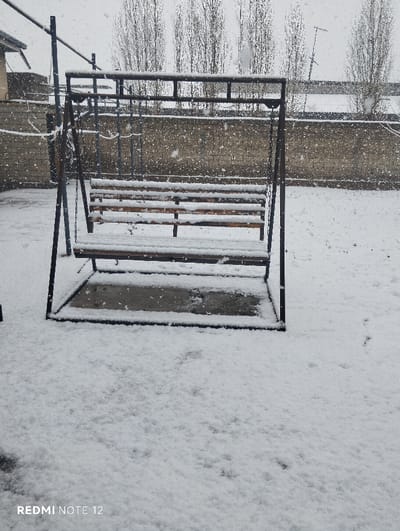 This image depicts an outdoor scene in Beshtam, Uzbekistan, during active snowfall. The foreground and mid-ground are entirely covered in a layer of fresh snow. A dark-colored metal-framed swing set with a wooden bench, also covered in snow, stands centrally. Faint tracks are visible in the snow on the ground, particularly in the lower right. In the background, a light-colored stone or concrete wall runs horizontally across the frame. Beyond the wall, several tall, bare deciduous trees are visible against an overcast sky. To the far left, a section of a building with a roofline is partially seen, and a metal pole structure extends from it. On the right, above the wall, a tall antenna is present. No people or animals are visible.