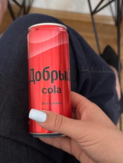 A person's hand is holding a red soda can. The can has text in Cyrillic and English. The Cyrillic text reads "Добры" which translates to "Dobry" and the English text reads "cola". Beneath the word "cola" is smaller text that reads "без сахара" which translates to "sugar-free". The person's fingernails are painted white. The background appears to be a dark blue fabric, possibly jeans. The lighting suggests it is daytime, indoors. The scene is a close-up of the hand and the can, with the background out of focus. The setting is not clearly identifiable beyond the fabric. No specific activity or event is taking place, other than the holding of the can. The overall impression is a casual, everyday moment.
