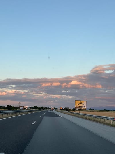 The image depicts a highway in the vicinity of Shtarkovo, Bulgaria, during sunset. The sky is a gradient of light blue to pink, with scattered clouds tinged with the warm hues of the setting sun. A billboard with the text "DEON SPORT ТВОЯТ УСПЕХ Е ВАЖЕН!" and a logo featuring a stylized animal is visible on the right side of the road. The highway is asphalt, with clear lane markings. Guardrails line the side of the road. In the distance, fields of dry grass and a line of trees are visible. There are no people or animals immediately apparent in the scene. The overall atmosphere is calm and serene.