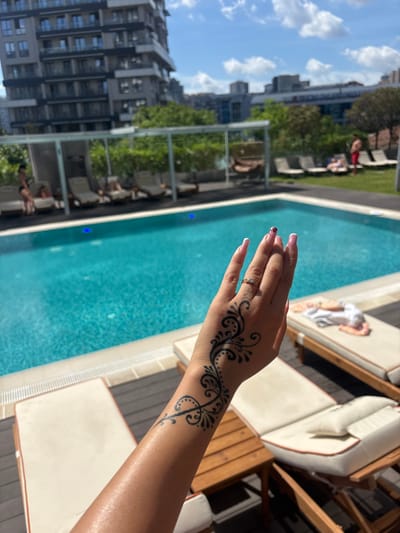 A person's arm, adorned with a black henna tattoo of swirling vines and dots, is the central focus of this image. The arm is extended towards a bright blue swimming pool, suggesting the activity of swimming or relaxing by the pool. The tattoo is intricate, covering the back of the hand and extending up the forearm. A ring with two small stones is visible on one of the fingers.

The background reveals a luxurious outdoor setting. A swimming pool with clear blue water occupies a significant portion of the frame, with what appear to be underwater lights creating subtle glows. Surrounding the pool are comfortable lounge chairs with white cushions and wooden frames, indicating a resort or a high-end residential area. In the distance, modern high-rise buildings rise against a partly cloudy sky, with lush green trees interspersed between them. The sunlight suggests it is daytime, likely a warm and pleasant day given the poolside setting. The overall mood is one of leisure and relaxation, enjoying a sunny day at a desirable location. The scene appears to be in Kâğıthane, Turkey, based on the provided context. No visible text is present in the image.