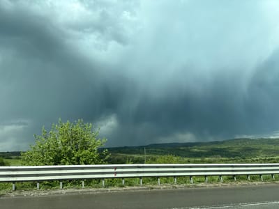 The image captures a landscape near Vakarel, Bulgaria, dominated by an approaching heavy storm. In the foreground, a section of a roadside barrier runs horizontally across the lower portion of the frame, bordering a paved road. Behind the barrier, a vibrant green bush stands out against the darkening sky. Beyond the bush and barrier, the land slopes gently, covered with lush green vegetation and dotted with trees, leading to distant hills. The sky is filled with thick, dark storm clouds, with a visible downpour occurring in the middle distance. The overall atmosphere is ominous, suggesting an imminent heavy rainfall.