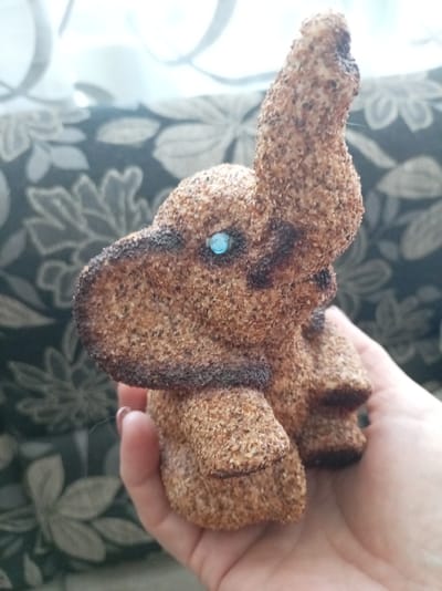 A small, textured elephant figurine, likely made of a material resembling sawdust or coarse sand, is held in a person's hand. The elephant has a raised trunk, a single turquoise or blue gem for an eye, and dark brown markings that define its ears, face, and feet. The figurine is positioned against a backdrop of a dark green fabric with a repeating floral or leaf pattern in a lighter shade. Above this fabric, a blurred white area suggests a window with curtains or blinds, allowing diffused light to enter. The overall lighting is soft and somewhat overcast, not indicating a specific time of day but rather a generally bright interior. The image has a shallow depth of field, with the elephant in sharp focus and the background elements blurred. The hand holding the figurine is that of an adult, with visible fingernails. The setting appears to be indoors, possibly a living room or bedroom. No visible text is present in the image. The emotional tone conveyed is neutral, focusing on the presentation of the object. The context of Bologoe, Russia, is provided externally and is not visually discernible from the image itself.