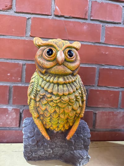 A close-up, eye-level shot shows a decorative ceramic owl figurine positioned in front of a red brick wall. The owl is depicted with its head tilted slightly to its left, displaying large, dark, reflective eyes and an orange-brown face with textured details resembling feathers. Its body is primarily a gradient of green and yellow, with intricate feather patterns painted onto its surface. The owl is perched on a dark gray, rocky base with a textured, rough finish. The brick wall behind the owl is composed of rectangular red bricks with visible gray mortar lines. The lighting appears to be ambient, casting subtle shadows. The scene is static and devoid of human presence or activity.