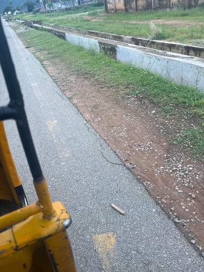 The image depicts an outdoor scene, likely taken from a vehicle, given the perspective and the presence of yellow and black railings in the foreground. The setting appears to be a roadside adjacent to a grassy embankment and a concrete structure, possibly a drainage channel or a retaining wall. The ground surface is a mix of asphalt with yellow markings and a dirt path scattered with small stones and pebbles. Sparse green grass grows along the edge of the embankment.
In the distance, on the left side of the frame, there are glimpses of buildings, suggesting an urban or semi-urban environment. The sky is not visible, but the lighting suggests it is daytime, possibly overcast or early morning/late afternoon. There are no people visible in the image.
A single, thin twig lies on the asphalt surface, a small detail in the overall scene. The image is oriented vertically, which may be due to the camera's orientation or the nature of the photograph. The overall impression is one of a functional, somewhat utilitarian environment, perhaps a construction site or an industrial area given the presence of heavy machinery implied by the yellow railings. The location context provided, Kona Garu, Nigeria, places the scene in a specific geographical region.