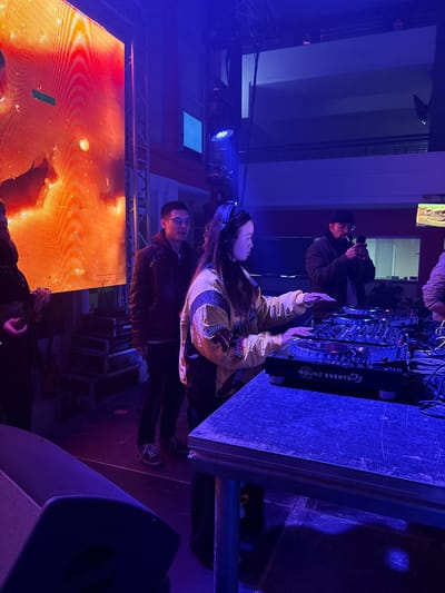 The image shows a woman in a yellow jacket with her hands near a DJ setup labeled "BOUNZ EVENTS DJ". A man in glasses stands beside her. A person in a black cap takes a photo in the background. A large screen displaying orange and yellow patterns is in the foreground. The scene is lit with blue stage lighting. Equipment and stage elements are visible, creating an atmosphere of an event.