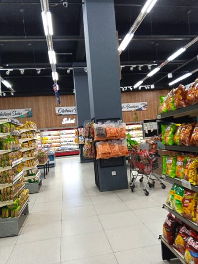 This media file presents an indoor view of a well-lit supermarket or grocery store aisle, likely situated in Sindanglaya, Indonesia. The perspective looks down a long, central aisle towards the rear of the store.
On the left side, tall shelving units are densely packed with various packaged goods, appearing to be snacks, instant noodles, or similar items. Opposite, on the right, more shelves display an array of colorful packaged snacks. Large, dark gray pillars are prominent in the middle of the aisle, with smaller bags of packaged items, possibly nuts or dried goods, hanging from hooks attached to them. A red shopping cart is partially visible on the right, positioned near the snack shelves.
The floor is covered with light-colored tiles, and the ceiling is black, adorned with bright fluorescent tube lights and track lighting fixtures, illuminating the entire space. In the distance, at the back of the store, refrigerated display cases or deli counters are visible along the wall, which is accented with wooden paneling above.
Visible text on signs along the back wall includes "Olahan Susu" (Processed Dairy) on the left, and further right, "Olahan Buah-Buahan" (Processed Fruits) with the word "Salat" (Salad) illuminated below it, indicating specific product sections. A small security camera is mounted on the black ceiling in the upper left portion of the image. The store appears modern, clean, and well-organized, with no people present, suggesting a quiet moment during operational hours.