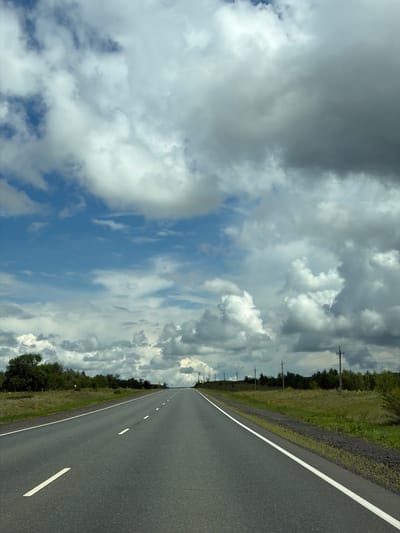 The image depicts an asphalt road stretching into the distance, flanked by grassy verges and a line of trees. The sky is filled with dramatic, dark clouds interspersed with patches of blue sky. A few telephone poles are visible along the roadside. There are no people or vehicles in the frame. The scene suggests a rural or highway setting. The location is the city of Ostrovnoye, Russia.