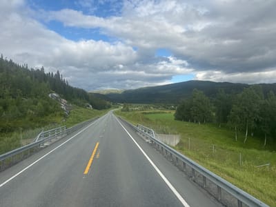 The media file depicts a view from the perspective of someone driving on a two-lane asphalt road in a rural, mountainous landscape. The road stretches into the distance, curving gently to the left. On the left side of the road, a steep, rocky embankment is covered with evergreen trees. On the right side, there is a grassy slope with scattered trees and a metal guardrail along the edge of the road. The sky is partly cloudy with dramatic cumulus clouds interspersed with patches of blue sky.
In the distance, rolling hills covered in dense forest create a layered backdrop. A small white car is visible further down the road. The overall impression is one of a scenic drive through a natural environment. The lighting suggests it is daytime, likely mid-afternoon, given the brightness and the shadows. The weather appears to be partly sunny with some clouds, typical of a summer day in Norway. There are no visible people or text in the image. The scene evokes a sense of peaceful travel and appreciation for natural beauty. The setting is consistent with the provided location context of Røssvoll, Norway, characterized by its mountainous terrain and extensive forests.