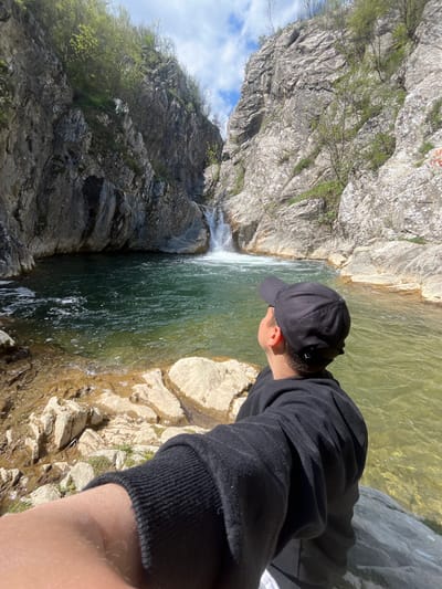 A man stands at the edge of a rocky cliff overlooking a waterfall, likely in the vicinity of Medven, Bulgaria. The man, wearing a black sweatshirt and a black baseball cap, is positioned on the right side of the image and is gazing down at the water below. The waterfall cascades from the top of the image, creating a pool of water surrounded by rocks. On the left side of the frame, there are tall cliffs with sparse vegetation. The sky is visible in the upper left corner, revealing a few clouds.