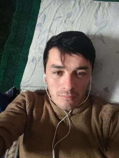 A close-up selfie shows a man with short, dark hair and brown eyes wearing a brown turtleneck sweater. He has fair skin and some stubble. White wired earphones are plugged into his ears, with the wires visible against his sweater. He is lying down, with his head resting on a patterned pillow. Part of a green textured surface is visible on the left side of the image. The lighting is soft and diffused.