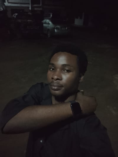 The image is a selfie taken at night, likely outdoors in a residential area of Orodo, Nigeria. The foreground is dominated by a young Black male with short hair and a light mustache, wearing a dark collared shirt. He is looking towards the camera with his head slightly tilted and is holding his left arm across his chest, with his right hand resting on his upper left arm. He is wearing a dark smartwatch on his left wrist.

In the background, out of focus, are several vehicles. One appears to be a pickup truck with "TOYOT" visible on its tailgate, suggesting it's a Toyota. To the right of the truck is a silver sedan. Behind the vehicles, there are dimly lit building structures, consistent with residential or commercial buildings. The overall lighting is dim, indicating it is nighttime, and the ground appears to be unpaved.