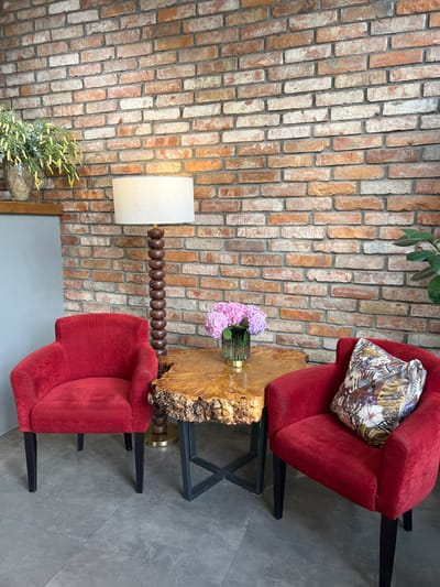 A scene is depicted in Adzhiryuara, Abkhazia, featuring two red armchairs positioned around a small, uniquely shaped wooden table. The table has a rough-edged, natural wood surface with a visible grain and a black metal base. On the table, a glass vase holds a bouquet of pink hydrangeas. A floor lamp with a white shade and a decorative wooden base stands between the chairs. The backdrop is a textured brick wall, exhibiting variations in brick color and grout. To the left of the arrangement, a shelf holds a vase with yellow-green foliage. To the right, part of a leafy plant is visible. One armchair has a decorative pillow with a floral and tropical leaf pattern. The floor appears to be tiled in a neutral gray. There are no people present in the image.