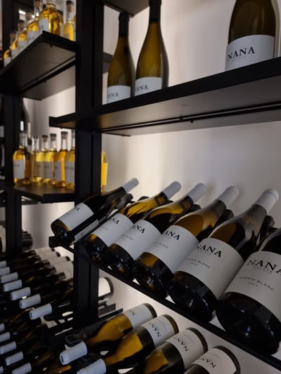 The image depicts wine bottles on racks, likely within a wine cellar or shop in Mizpe Ramon, Israel, as suggested by the "NANA ESTATE" labeling on the bottles. The bottles are arranged on black metal racks, offering a glimpse of multiple rows. Labels reveal varieties such as "CHENIN BLANC" and "CHARDONNAY" with some marked for the year "2025." The image is taken from a low angle, looking upwards, emphasizing the stacked and angled arrangement of the bottles. The lighting is soft, suggesting an indoor environment.