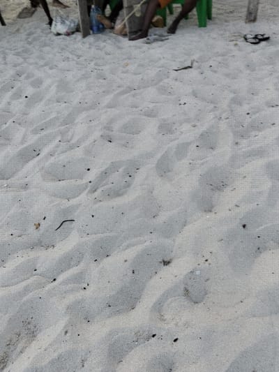 The scene depicts a sandy outdoor environment in Gombato Bongwe ward, Kenya. The foreground is dominated by light-colored sand exhibiting numerous indentations, consistent with footprints, and scattered small dark debris including minor organic matter.

In the upper portion of the image, the lower extremities of several individuals are visible. One person is seated in a green plastic chair, wearing orange shorts, with bare feet on the sand. Another individual's bare, dark-skinned feet are visible adjacent to a second green plastic chair. Additional dark-skinned legs and feet are partially observed towards the upper-left. A pair of black flip-flops rests on the sand to the upper-right.

Various objects are present on the sandy ground, including a white plastic bag, a blue plastic bottle, and other indistinct items. Vertical wooden posts, one secured with a light-colored rope, are also part of the background elements. No specific actions or direct interactions between individuals are discernible; the individuals appear to be resting or gathered. The lighting suggests ambient daylight.