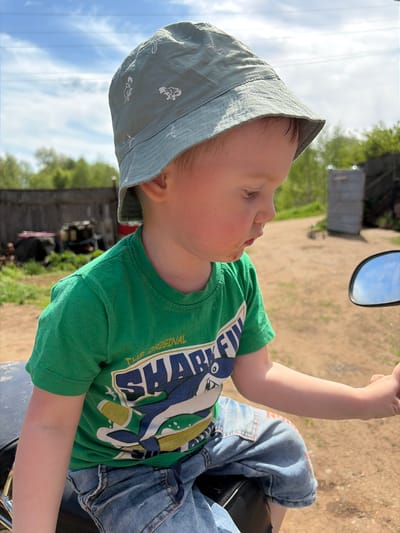 A young child sits on a motorcycle, wearing a light blue hat with dinosaur prints, a green "Shark" t-shirt, and denim shorts. The motorcycle has a side mirror visible. The background shows a dirt road, a wooden shed, and vegetation. The location is Ul'yanovka, Russia.