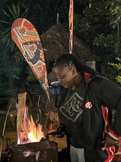 This media file captures an outdoor night scene in what appears to be Mururiini, Kenya. The image prominently features a person in the foreground, facing downwards and to the left of the frame. They have dark skin and dark, braided hair with small gold earrings visible. They are wearing a black jacket with red and white trim, featuring a distinctive white graphic design on the left side of the chest.
In the lower right portion of the image, a robust fire burns brightly within a metal brazier or fire pit, casting light on its surroundings. Flames and sparks are visibly rising from the fire, suggesting warmth, cooking, or a communal gathering. Several logs or pieces of wood are arranged around the fire, fueling it.
A large, paddle-shaped banner or sign stands in the mid-ground, angled towards the left. It features vibrant patterns in red, orange, and black, and displays repeated text: "CUCURUCHA RUI MUKARU". Accompanying the text are cartoon-like illustrations of a figure with a bowl on their head. The background is dark, indicating it is nighttime, with silhouettes of green foliage visible. The overall setting suggests a casual, possibly festive, outdoor event or gathering, potentially involving food or community celebration.