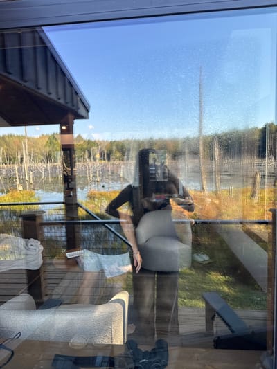 An image captured through a glass window reveals both interior reflections and an exterior landscape in Otego, United States. The exterior scene depicts a body of water, likely a pond or swamp, with numerous bare tree trunks emerging from the water. Behind the water, a forested area with autumn foliage extends to the horizon under a clear blue sky. A deck railing is visible in the mid-ground. The window pane itself is speckled with droplets or debris.
Reflected in the window, a person, appearing to be an adult female with long, dark hair and dark clothing, stands holding a small, light-colored object (possibly a phone or camera) in her right hand, positioned as if capturing the scene. To her right, a light-colored, upholstered armless chair is reflected, and to her left, a stack of white towels rests on a surface. Below these, partial reflections of another upholstered chair, a folded dark chaise lounge or beach chair, and a pair of light-colored shoes are visible on what appears to be an exterior deck surface. On the interior wooden floor, reflected at the bottom, a pair of binoculars and a dark controller (possibly a gaming controller) are present.