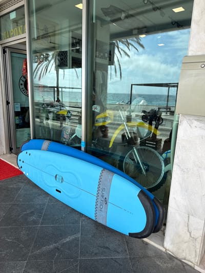 This is a photograph taken at eye level, looking slightly upwards. The main subjects are two light blue surfboards propped up against a building, with their tails on the ground. They appear to be new and unused. Behind the surfboards, a large glass window reflects the scene. Through the glass, we can see a yellow motorcycle, a bicycle wheel, and further in the distance, the ocean. The scene is set outdoors, with a stone-tiled pavement in the foreground. The reflection in the window shows a bright, overcast sky with palm trees visible. There is a sign on the surfboard that reads "SURFTECH" and another logo with text that is difficult to read. Another sign on the window reflects text that appears to say "JAPONA COLL". The overall impression is that of a surf shop in a coastal town, possibly in Sesimbra, Portugal, given the location context. The lighting suggests it is daytime, with diffused light indicating an overcast or cloudy day. There are no people visible in the image, and the focus is on the objects and the reflected environment.