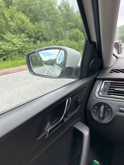 The image shows the interior of a car, viewed from the passenger side. The car is in motion on a paved road, with lush green trees visible on either side of the road and reflected in the side mirror. The side mirror, slightly dirty, shows a reflection of the road ahead and the sky.
Inside the car, the black door panel is visible, with a chrome door handle. To the right, part of the dashboard is in view, including air vents with chrome trim, and controls for lights and window adjustment. A green bottle cap is visible at the bottom of the frame, suggesting the presence of a bottle.
The sky is cloudy with patches of blue. There are some water droplets on the car window and mirror, indicating recent rain or humidity. The overall impression is a journey through a green, natural environment.
The location is the city of Arkhangelskoye, Russia.