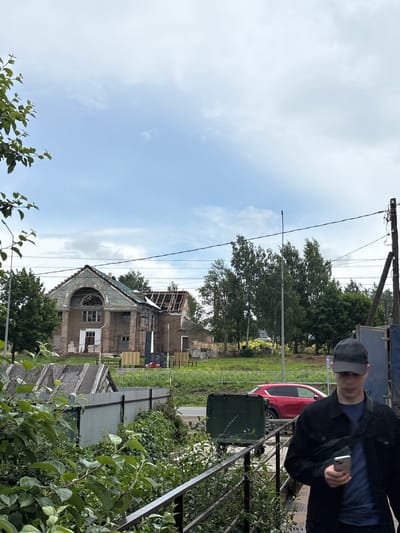 The image depicts a street scene in the city of Kstinin, Russia, under an overcast sky. In the mid-ground, a dilapidated building, possibly undergoing renovation or demolition, is visible. The structure has a damaged roof with exposed rafters and a large arched opening on its facade. Trees and foliage surround the building, and a road runs in front of it. A red car is parked on the side of the road. In the foreground, a young man wearing a black baseball cap and a black jacket is walking, holding a smartphone in his right hand. He appears to be looking at the phone. Various elements of urban infrastructure, such as a metal fence, a lamppost, and power lines, are present in the scene. The overall impression is one of urban decay and ongoing construction or deconstruction.