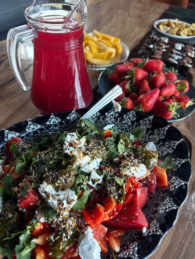 The image shows a table with a variety of food items. There is a black ornate bowl filled with a salad composed of tomatoes, red peppers, leafy greens (possibly cilantro and basil), and dollops of what appears to be fresh cheese, topped with mixed seeds and green dressing. To the side, there's a pitcher of red liquid, likely a juice or beverage. Further back, there are plates of strawberries, cut mangoes, and what could be chocolate pieces. The scene gives the impression of a prepared meal or snack spread. There is no information available to suggest a location of Дубровка, Russia.