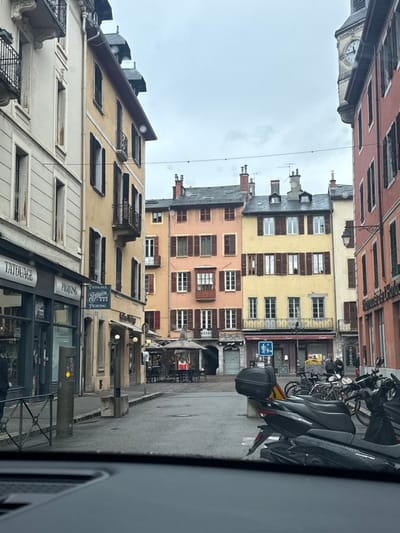 This image depicts a street scene in Chambéry, France, specifically around the vicinity of Place Saint-Léger, looking towards the Tour de l'Horloge in the background. The photograph is taken from inside a vehicle, with the dashboard or windshield frame visible at the bottom.
The scene features a wet, paved street or pedestrian square, indicating recent precipitation. The sky is overcast and uniformly grey.
On the left side, multi-story historic buildings line the street. The leftmost visible building is cream-colored, while the adjacent one is a pale yellow/ochre. Both feature multiple windows with dark, closed shutters and some wrought-iron balconies. At street level, two commercial establishments are visible: a "TATOUAGE" (tattoo) parlor and a "PIERCING" studio, with the prominent sign "TATOUAGE STUPIDIN 777 PIERCING." A dark grey bollard or payment terminal with a small yellow light is present on the sidewalk. A person in dark clothing is partially visible near the tattoo parlor entrance.
In the center-midground, the street widens into a square flanked by a row of colorful, multi-story buildings. From left to right, these include a pink/terracotta building with dark brown shutters and a distinctive enclosed wooden balcony on its second floor, followed by a yellow building with similar shutters and a wrought-iron balcony. An archway or passage is visible at ground level beneath the pink building. In front of these buildings, an outdoor seating area for a café or restaurant is set up with tables, chairs, and several light-colored, large umbrellas. A blue pedestrian/bicycle sign is mounted on a pole nearby. A few indistinct figures are seated at the outdoor tables, and two additional figures are walking further right near parked vehicles.
On the right side of the image, another multi-story building with a reddish/orange facade and dark shutters is present. A black, ornate street lamp extends from this building. Parked motorcycles and bicycles line this side of the street; the closest motorcycle is dark-colored, possibly a scooter, equipped with a black top case.
In the upper-right background, a prominent, light-colored stone clock tower, likely the Tour de l'Horloge, rises above the rooftops. Its clock face is visible, indicating a time of approximately 11:40. Thin overhead wires traverse the sky across the street.