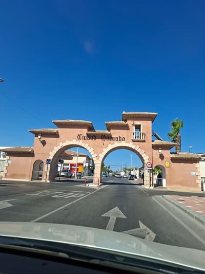 The image shows an archway in Rojales, Spain, that marks the entrance to Ciudad Quesada. The arch is made of a reddish-brown material and has "CIUDAD QUESADA" written in black lettering across the top. The arch spans a road with clear lane markings and traffic signs. There are cars on the road, and pedestrians are visible on the sidewalks. The sky is clear and blue, indicating a sunny day. The architecture of the archway suggests a Spanish colonial style, with tiled roofs and decorative stonework. The overall impression is of a well-maintained urban entrance.
