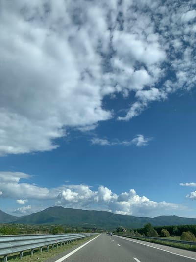 This daytime photograph captures a scenic view from a vehicle on a highway, with the image oriented vertically to the left. The scene is dominated by a bright blue sky filled with scattered white cumulus clouds, ranging from wispy formations to more substantial, fluffy clusters. In the distance, a majestic range of green mountains runs parallel to the road, with some peaks partially obscured by larger clouds. Sparse trees are visible on the lower slopes and closer to the highway.
On the right side of the frame, a multi-lane highway stretches into the distance, marked with clear white lines defining lanes and the shoulder. A metallic guardrail separates the road from the adjacent landscape. Further along the road, a few dark-colored vehicles can be seen, appearing small due to the distance, indicating traffic moving in the same direction as the photographer. High on the mountain ridges, faint shapes suggest the presence of wind turbines. The overall impression is one of open spaces and travel through a mountainous region under a clear, bright sky, likely in Marino pole, Bulgaria. There is no visible text.
