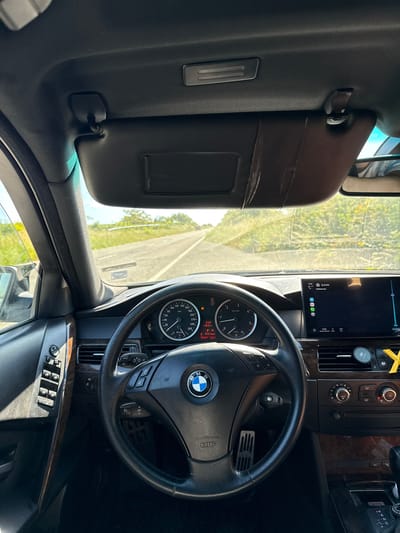The image shows the interior of a BMW car, focusing on the driver's side. The dashboard, steering wheel with the BMW logo, instrument panel, and center console are visible. The car is equipped with a navigation screen showing a map. The speedometer indicates the car is not in motion. The sun visor is lowered on the driver's side. The navigation screen suggests a location in or near Izvorishte, Bulgaria. The road outside the car is visible through the windshield.