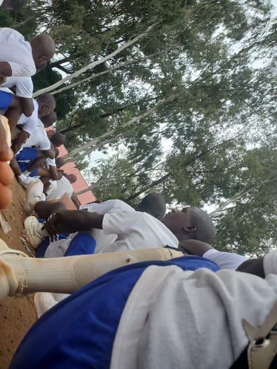The image depicts a group of young men, seemingly in a training or exercise setting, outdoors. They are dressed in white t-shirts and blue shorts or athletic pants. Some are sitting on the ground, others are in a resting or stretching pose. The environment appears to be a dusty or sandy area, with trees and foliage in the background, suggesting a rural or semi-urban location. The overall scene conveys a sense of physical activity and camaraderie. The text references "Bahit, Nigeria" as the location, implying this is a specific outdoor area within that Nigerian city.