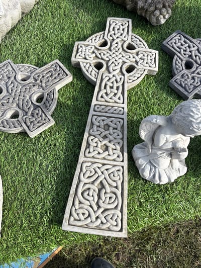 The image shows a collection of stone objects resting on artificial green turf. Prominently featured are three Celtic crosses, each with intricate knotwork designs. A small, weathered statue of an angel is also present. The objects appear to be displayed for sale, possibly in an outdoor setting in Chirk, United Kingdom. The lighting suggests a sunny day, with visible shadows cast by the objects. The composition is a high-angle shot, giving a clear view of the items' details and placement.