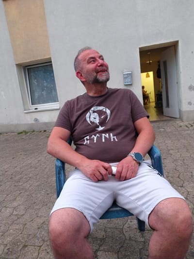 A light-skinned, bearded man with graying hair sits in a blue plastic chair outdoors. He is wearing a brown t-shirt with a white graphic and the word "SYNIA" in white lettering, and light gray shorts. He has a watch on his left wrist. The man is looking up and to his right with a slight smile. The background consists of a building with a light-colored exterior wall and a window, and a doorway that leads to an interior space with a person sitting. The ground is paved with rough, interlocking stones. The overall scene appears to be in a residential or commercial outdoor area in the city of Herford, Germany, possibly near a private residence or a small business.